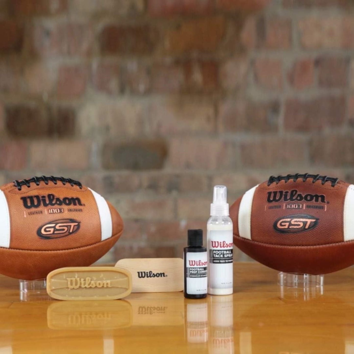 Two Wilson GST footballs with cleaning products on a reflective surface against a brick wall.